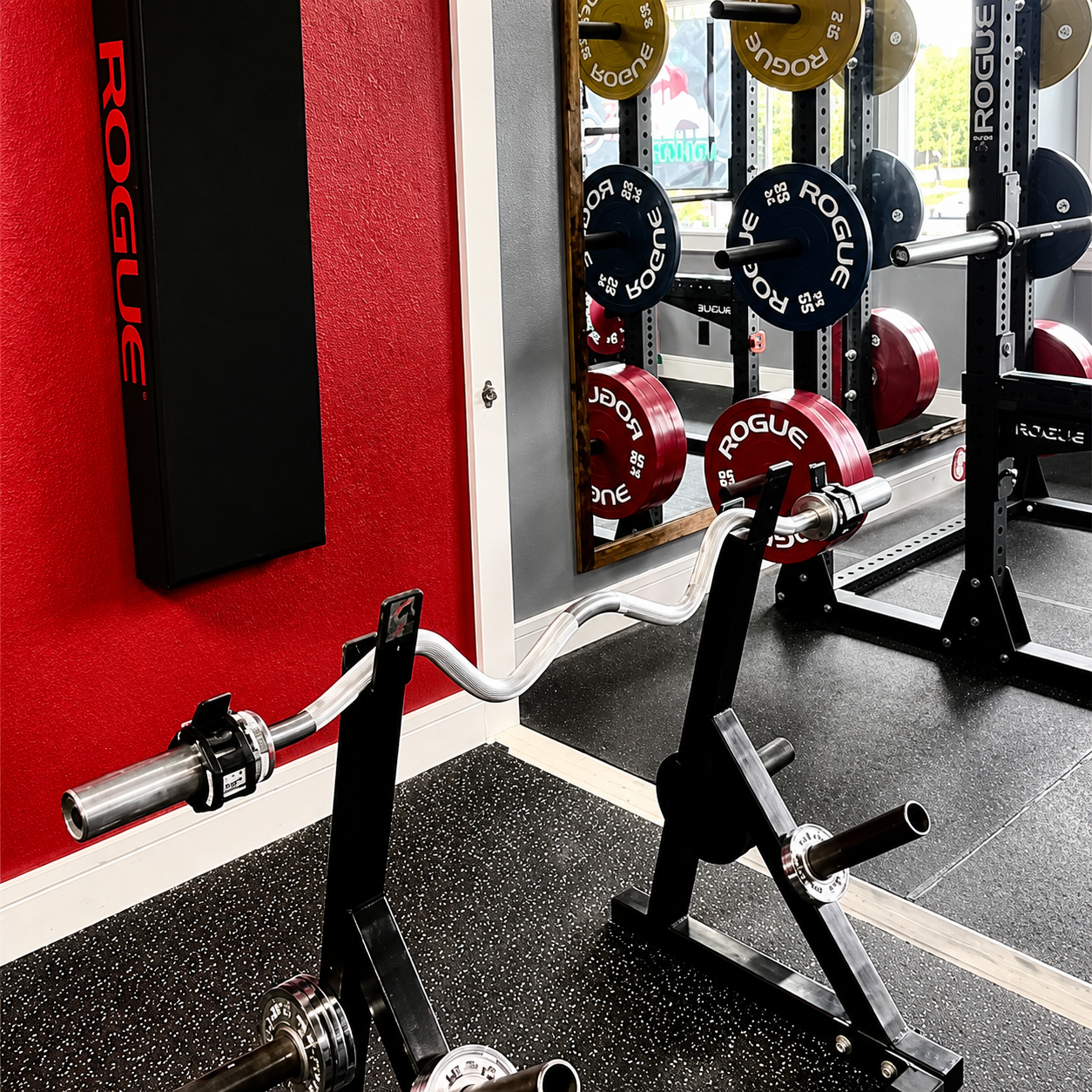 An EZ curl bar resting on stands against a red wall, with loaded racks reflected in a mirror