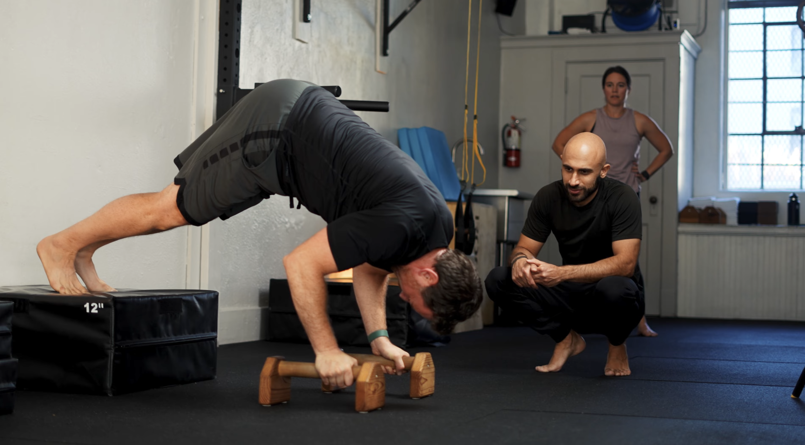 Client performing an elevated pike press on parallettes while a coach watches closely at Capra Bodyweight Training