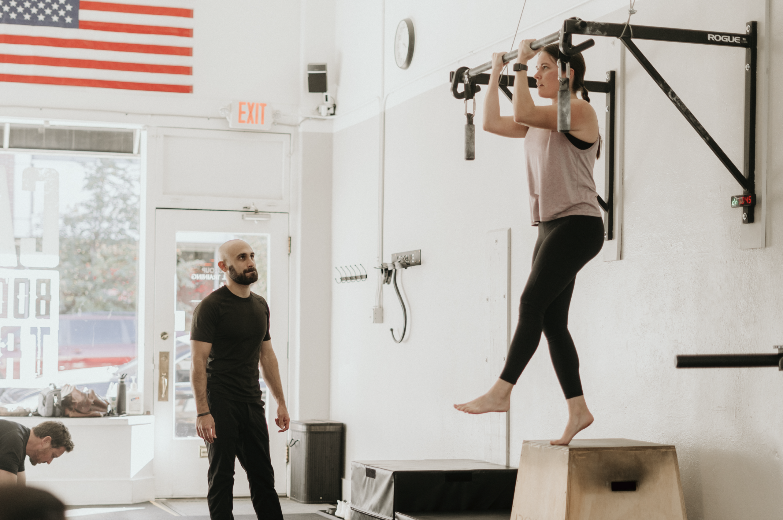 Client starting a pull-up from an elevated box while a coach observes at Capra Bodyweight Training