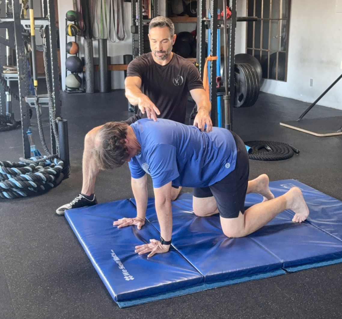 Aaron Delgrolice leans over a client on all fours, pointing along the client’s upper back during a mat drill