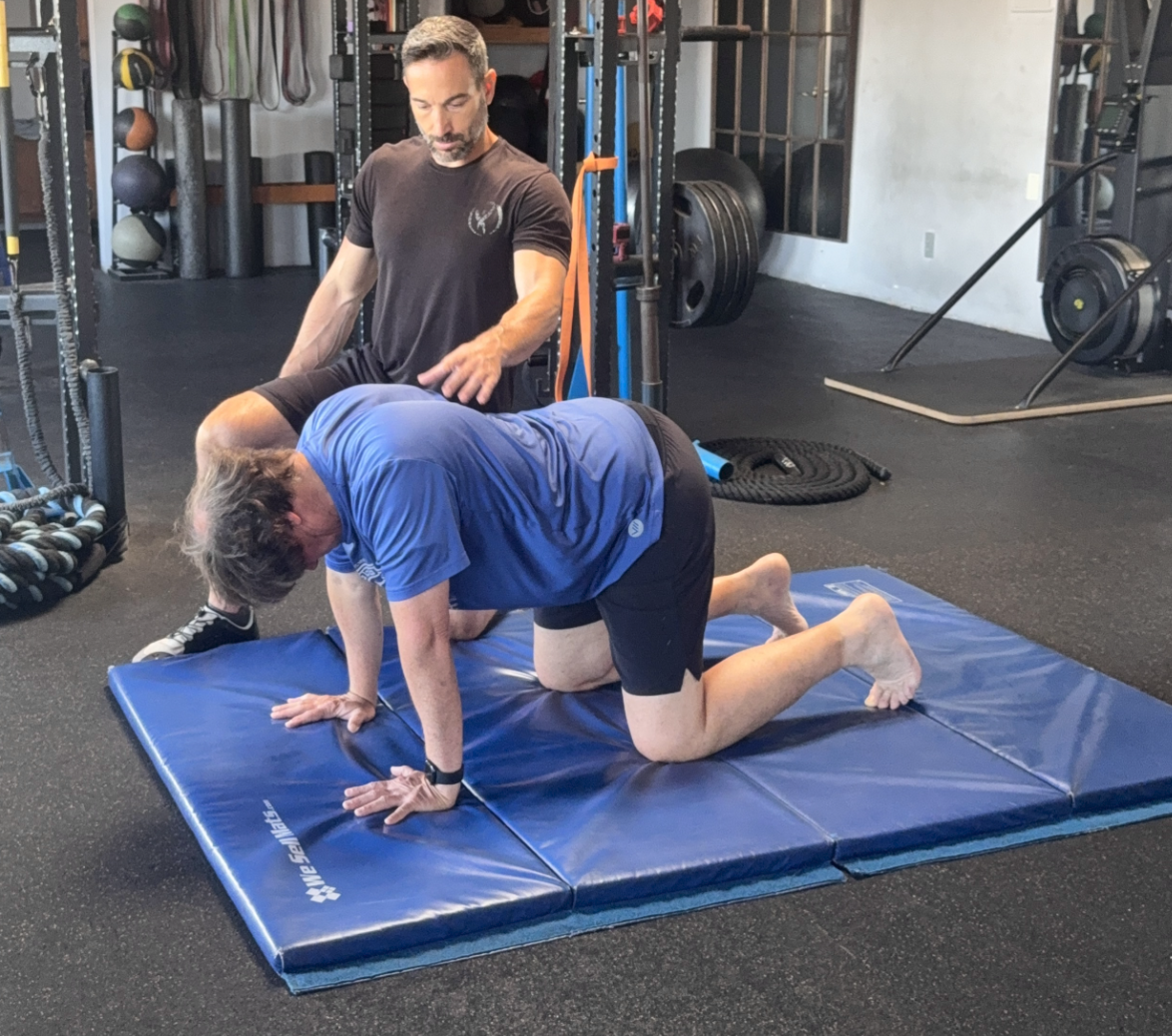 Aaron Delgrolice kneels behind a client on all fours on a blue folding mat, one hand on the client’s back