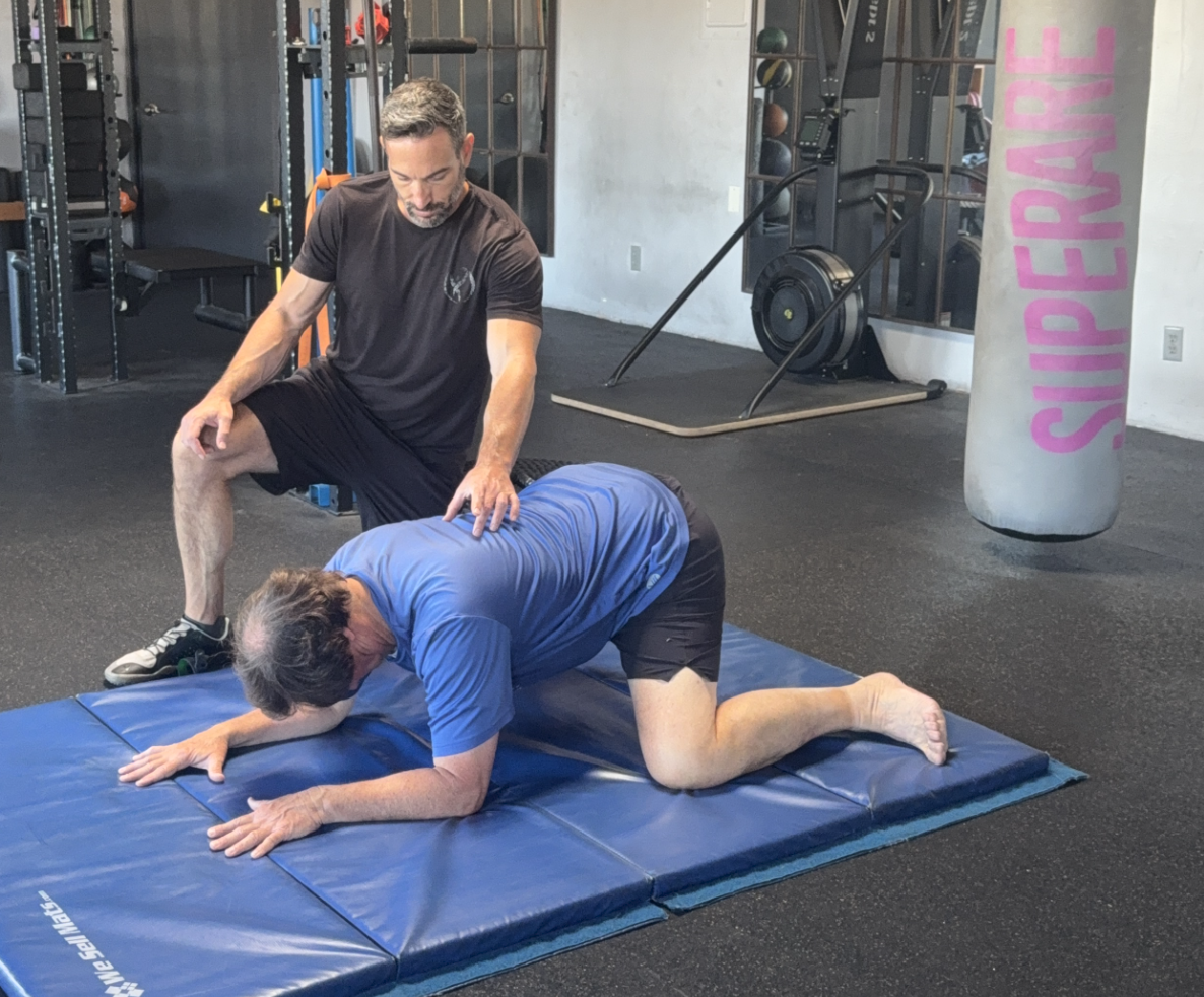 A coach kneels beside a client rocking back from all fours on blue mats, a heavy bag hanging nearby