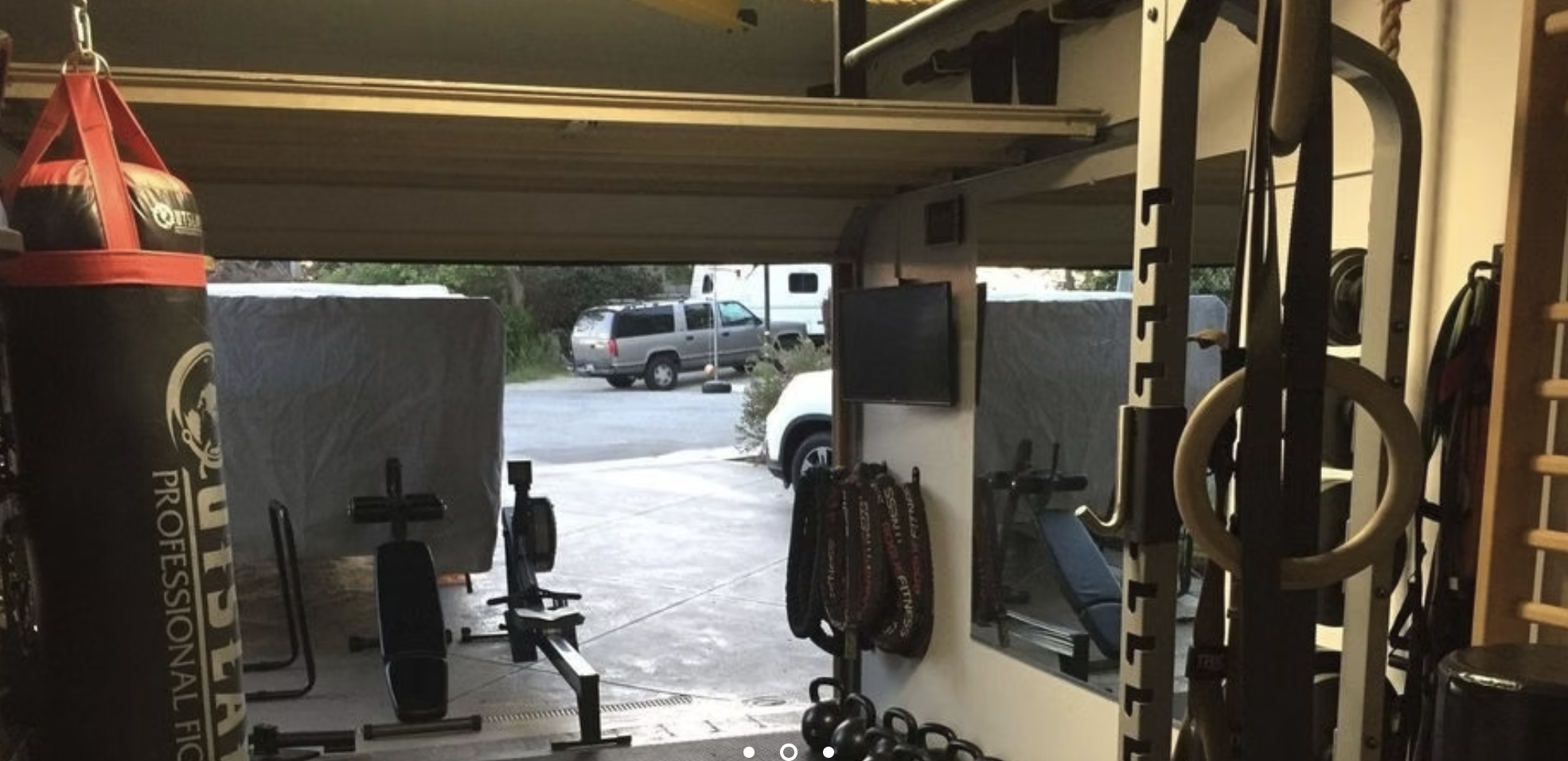 Small Box Fitness training room with the garage door open, showing a heavy bag, rowing machine, benches, kettlebells, and rack uprights