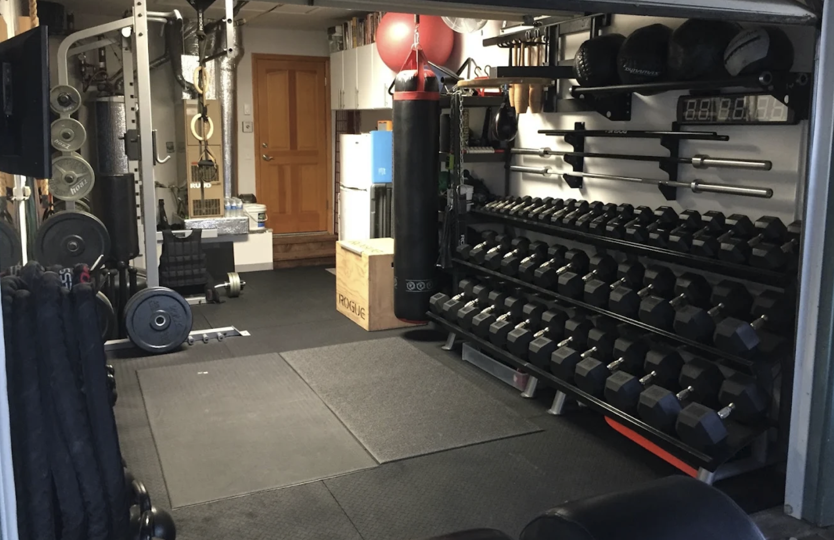 Interior of the Small Box Fitness training room with dumbbell racks, barbells, heavy bag, gymnastic rings, and matted floor