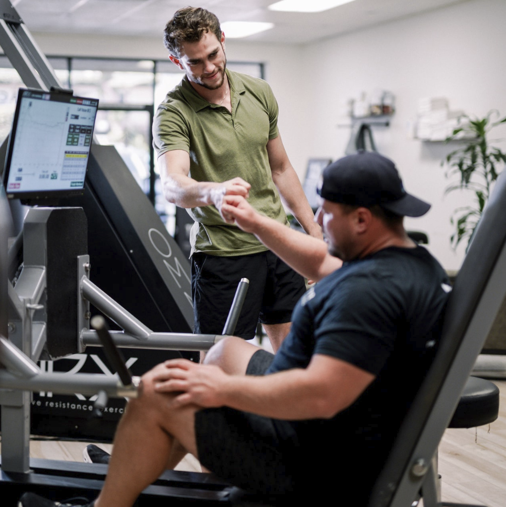 A coach fist-bumping a client seated at the ARX leg press at Top Tyr Training, with set output on the monitor