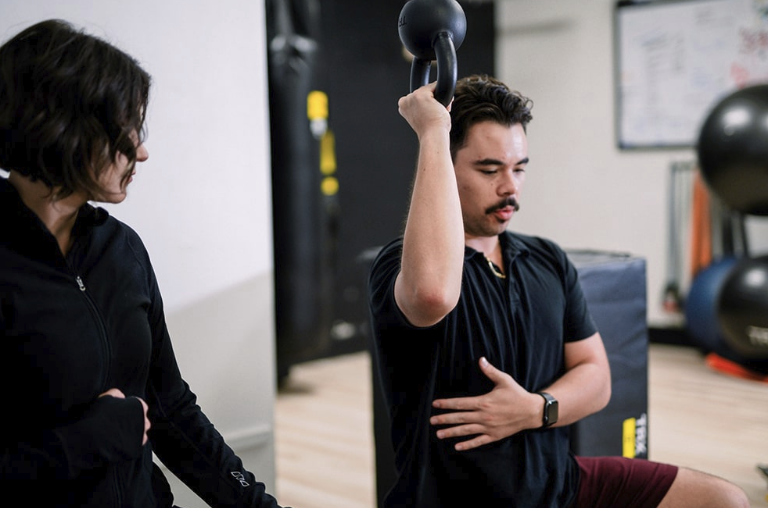 A coach watching an overhead kettlebell hold with a hand braced at the midsection at Top Tyr Training
